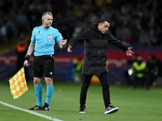 On the pitch, a football referee and Barca's coach Xavi discuss a play, with Xavi gesturing animatedly, arms outstretched.