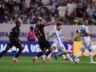 Soccer player in white and blue striped uniform dribbles the ball while three opposing players in dark uniforms and one teammate in the background run on a green field during an Auto Draft match.