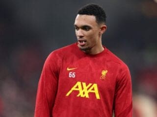 A Liverpool defender in a red football jacket stands on the field, glancing to the side.